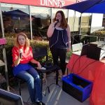 Two women sing karaoke under umbrellas at Augie’s, one seated, one by equipment.