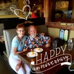 An older couple smiles at a Steakhouse Prescott booth with cake; text reads Happy anniversary.