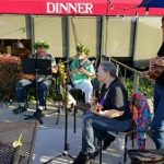 Four musicians play guitars and saxophone outside a Steakhouse Prescott under a red awning.