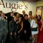 A group dressed formally poses by a wall sign reading Augie’s Cocktail Lounge in Prescott.