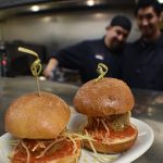 Two meatball sliders on a plate at Augie’s, with two smiling chefs in the restaurant kitchen.