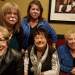 Five smiling women with fake mustaches pose indoors, ready for fun at Banquet Rooms Prescott.