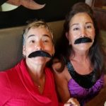 Two women smiling in fake mustaches at Augie’s, sitting together and having a laugh indoors.