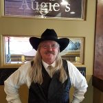 Man with long hair, black hat, glasses, and vest stands by an Augies sign—Restaurants in Prescott.