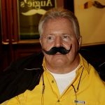 Man in a yellow shirt with a large fake mustache, indoors at restaurants in Prescott.