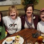Four friends with fake mustaches enjoy laughs at a Steakhouse Prescott restaurant table.