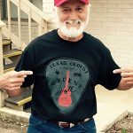 Smiling man in a red cap points to his Augie’s guitar T-shirt with a Gruene logo.