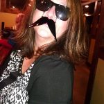 Woman at Augie’s in sunglasses and a fake mustache, posing indoors with a playful expression.
