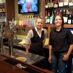Two women stand behind a bar at Steakhouse Prescott, smiling with bottles and taps on display.