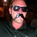 Man wearing sunglasses and a fake moustache, smiling indoors, dreams of Augie’s Steakhouse Prescott.