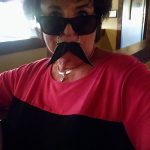 Person in sunglasses and fake mustache at Augie’s Cocktail Lounge, wearing a red and black shirt indoors.
