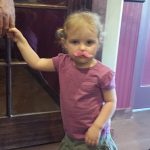 Young child with blonde hair in pigtails at Augie’s, wearing a pink mustache and purple shirt.