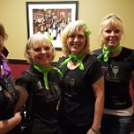 Four women at Augie’s, in black shirts and green scarves, smile together inside the restaurant.