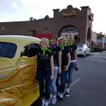 Four women with green scarves stand by a yellow vintage car outside Augie’s Cocktail Lounge.