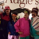 Four friends in retro 80s outfits and fake mustaches pose by a mural at Augie’s Cocktail Lounge.