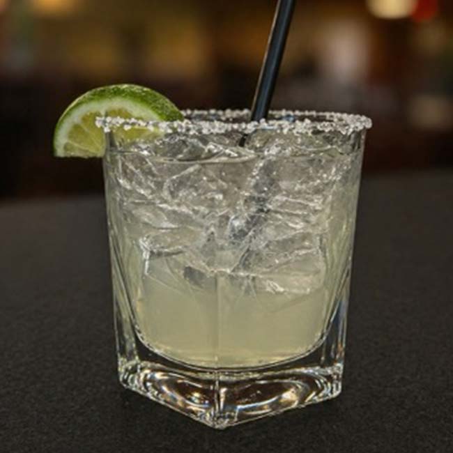 A margarita with ice, a salted rim, lime wedge at Augie’s Cocktail Lounge on a dark surface.