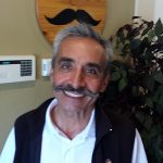 Smiling man with a mustache stands indoors near a plant at a Steakhouse Prescott location.