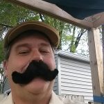 Man outdoors in tan cap, steakhouse Prescott vibe, with wooden structure and trees behind him.